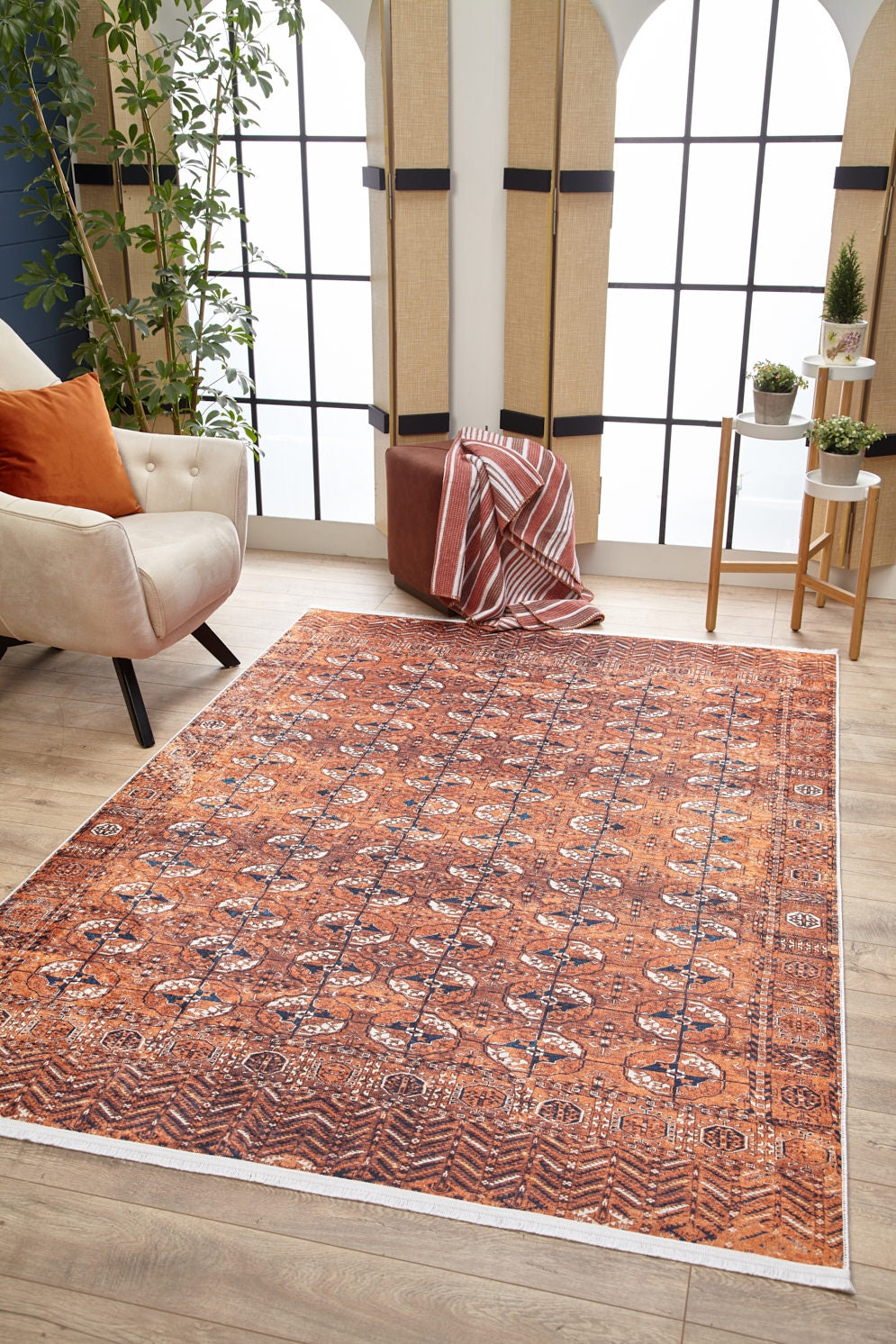 A living room featuring the Earthy Enigma - Washable Rug - JR1937 in orange, ideal for high-traffic areas, a white armchair with orange pillows, pouf with striped throw, plant stand, large windows, and tall corner plants.