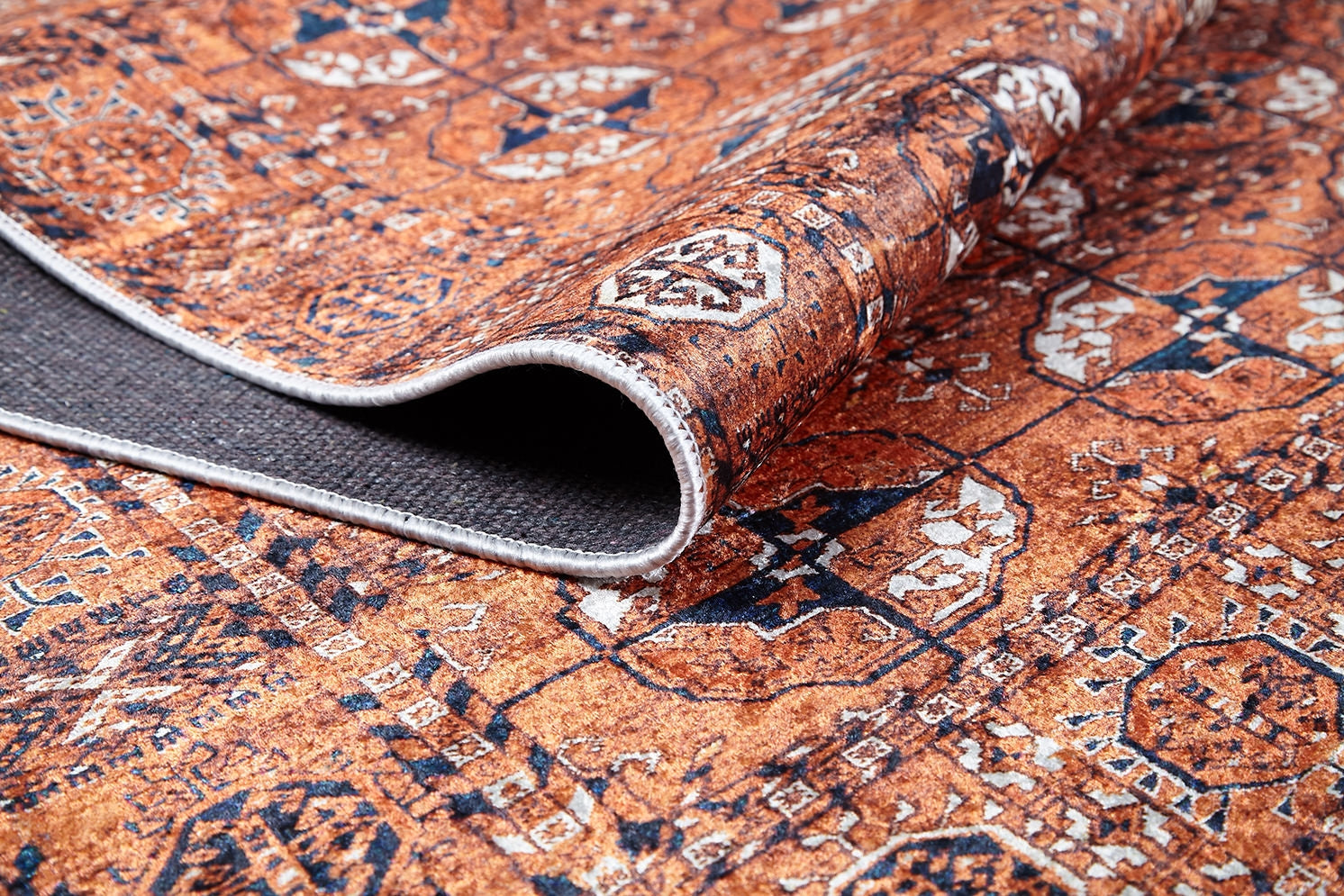 A close-up of the Earthy Enigma - Washable Rug - JR1937 shows its intricate rust-colored pattern; one edge curls to reveal the dark non-slip backing—ideal for high-traffic areas.