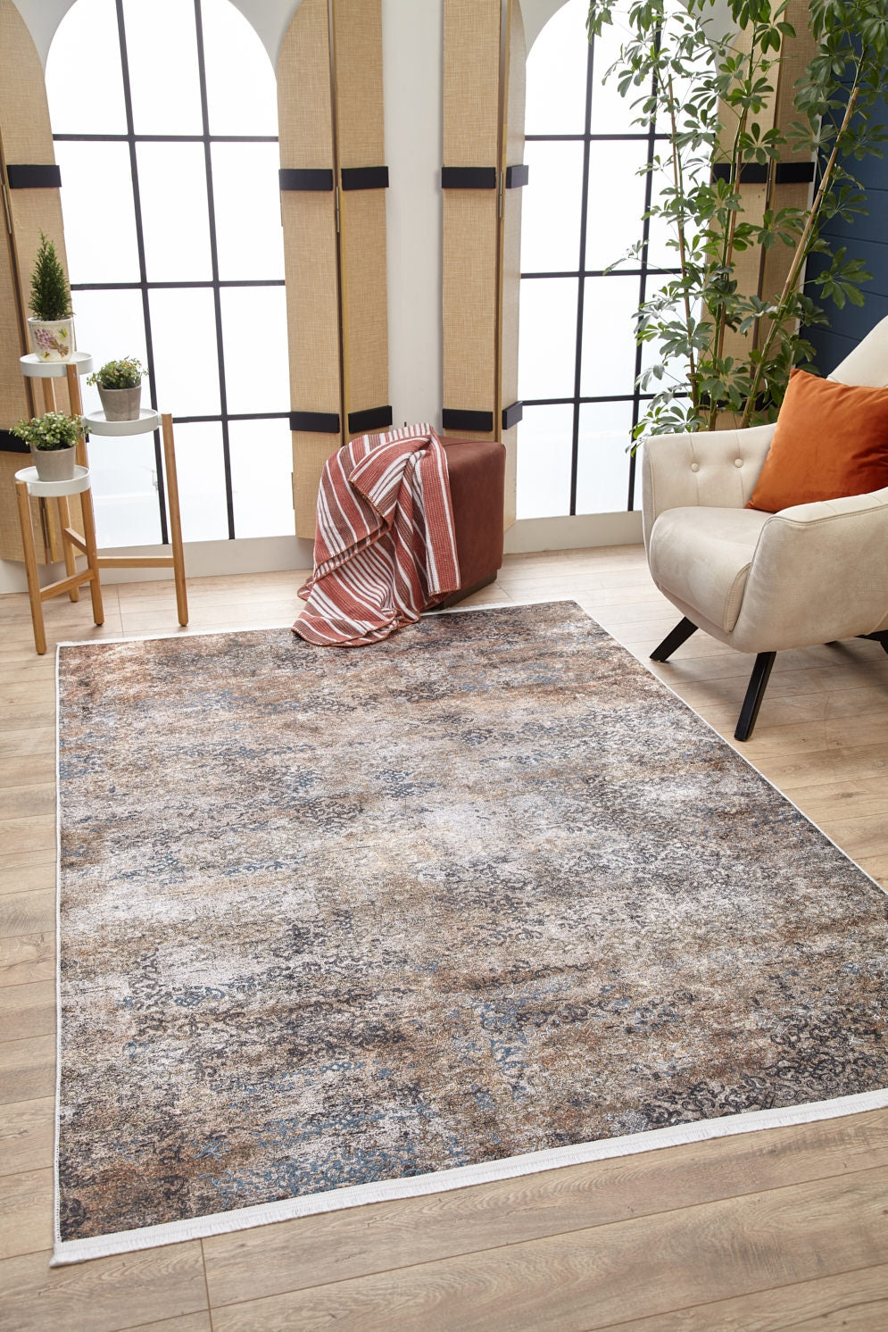 A modern living room features the Elegant Leaf - Washable Rug - JR1945 in bronze and brown on wooden floors, a beige armchair with an orange pillow, potted plant, side table with small plants, and soft light—perfect for high-traffic areas.