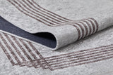 A close-up of the Elegant and Chic - Washable Rug - JR1509 shows its light gray surface with brown border lines and a lifted corner revealing a dark, textured underside—ideal for high-traffic spaces.