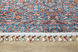 A close-up of the Ethereal Essence - Washable Rug - JR5087, a colorful flat weave with intricate blue, red, and orange geometric and floral designs, plus white fringe tassels with red and blue accents, shown on a wooden floor.