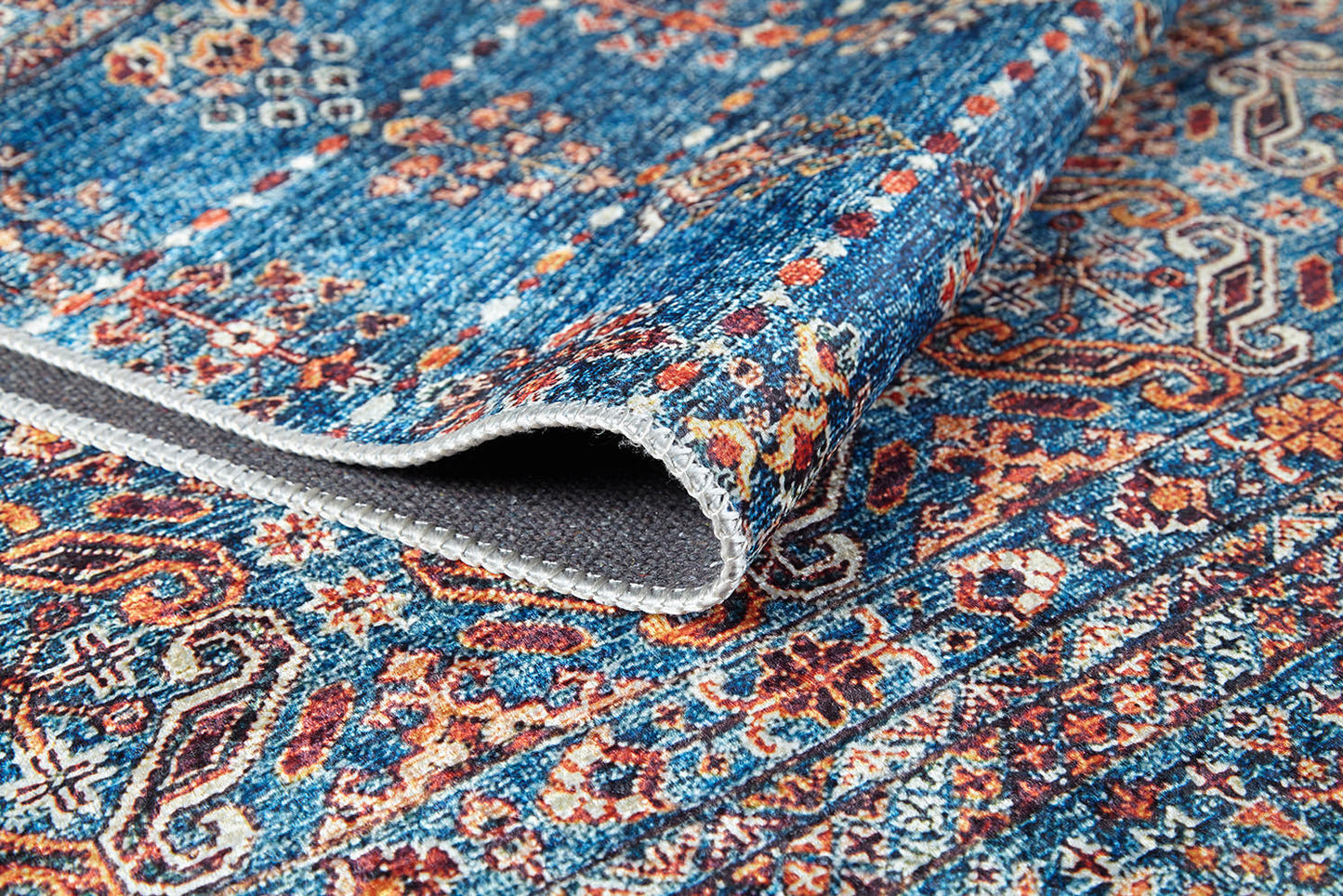 A close-up of the Ethereal Essence - Washable Rug - JR5087, folded to display its blue and orange flat weave, intricate floral and geometric patterns, textured washable fabric, and detailed edge stitching.