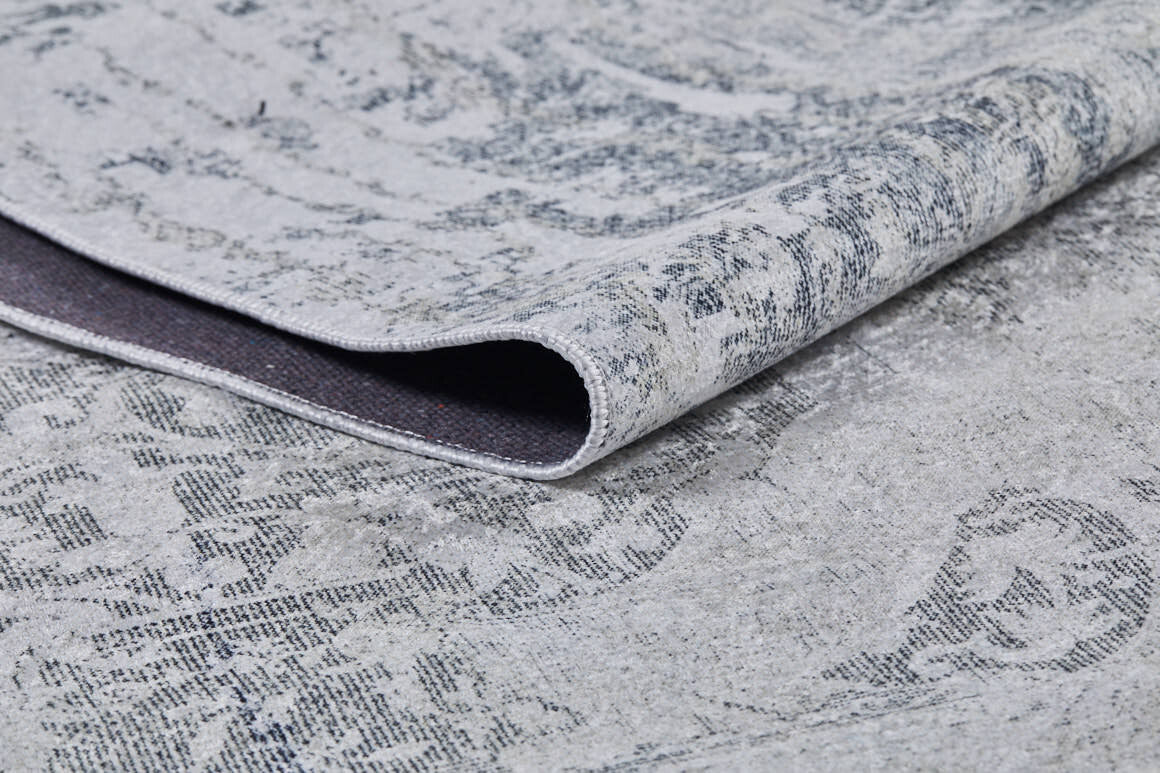 A close-up of the Faint Memories - Washable Rug - JR1437 in gray-anthracite, featuring a folded corner that shows its darker underside and slightly distressed, intricate surface design.
