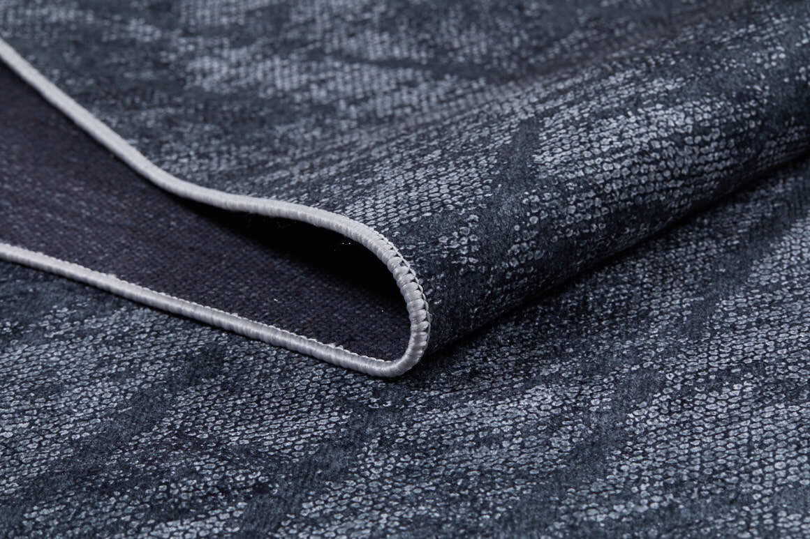 A close-up of the Geometric Gems - Washable Rug - JR1342 reveals its folded, textured dark gray fabric with a subtle pattern and light stitching—ideal for high-traffic areas due to its washable design.