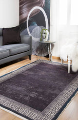 A modern living room with the Geometric and Chic - Washable Rug - JR1273 in dark gray with a Greek key border, a gray sofa, black pillow, white chair with fur throw, face vase with greenery on a table, and abstract wall art. Great for high-traffic areas.
