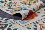 A close-up of the Global Gathering - Washable Rug - JR608 shows its multicolor floral and geometric design in red, blue, yellow, green, and black on a light background, with the corner lifted to reveal its textured underside.