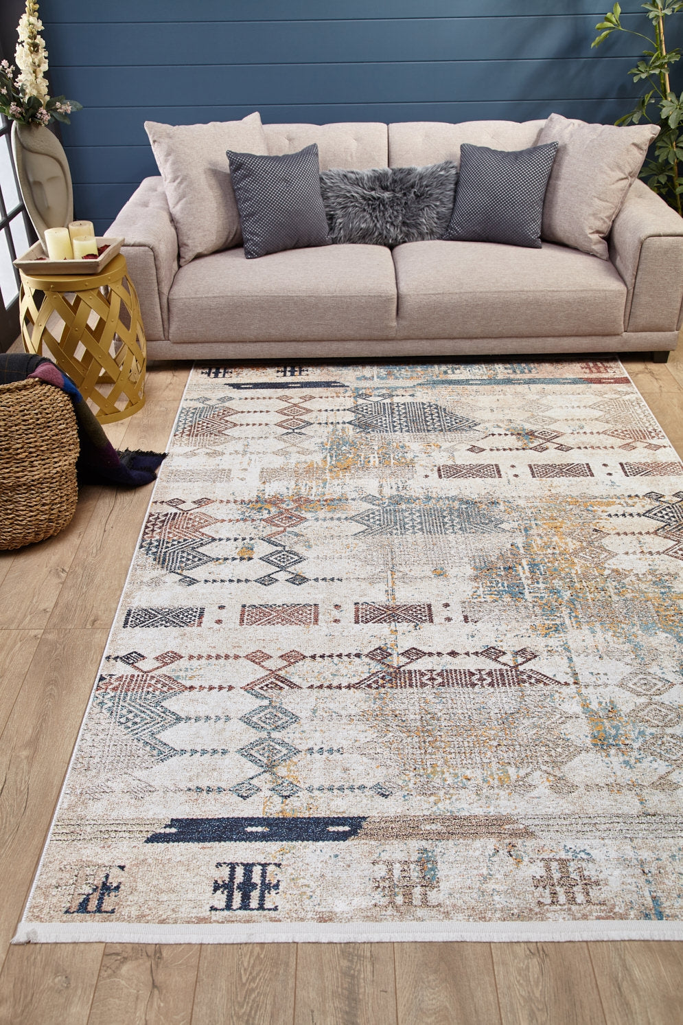 A modern living room features a beige sofa, gray and fur pillows, candles on a yellow side table, a woven basket, a plant in the corner on wood flooring, and the Global Glamour - Washable Rug - JR1708 with geometric designs.