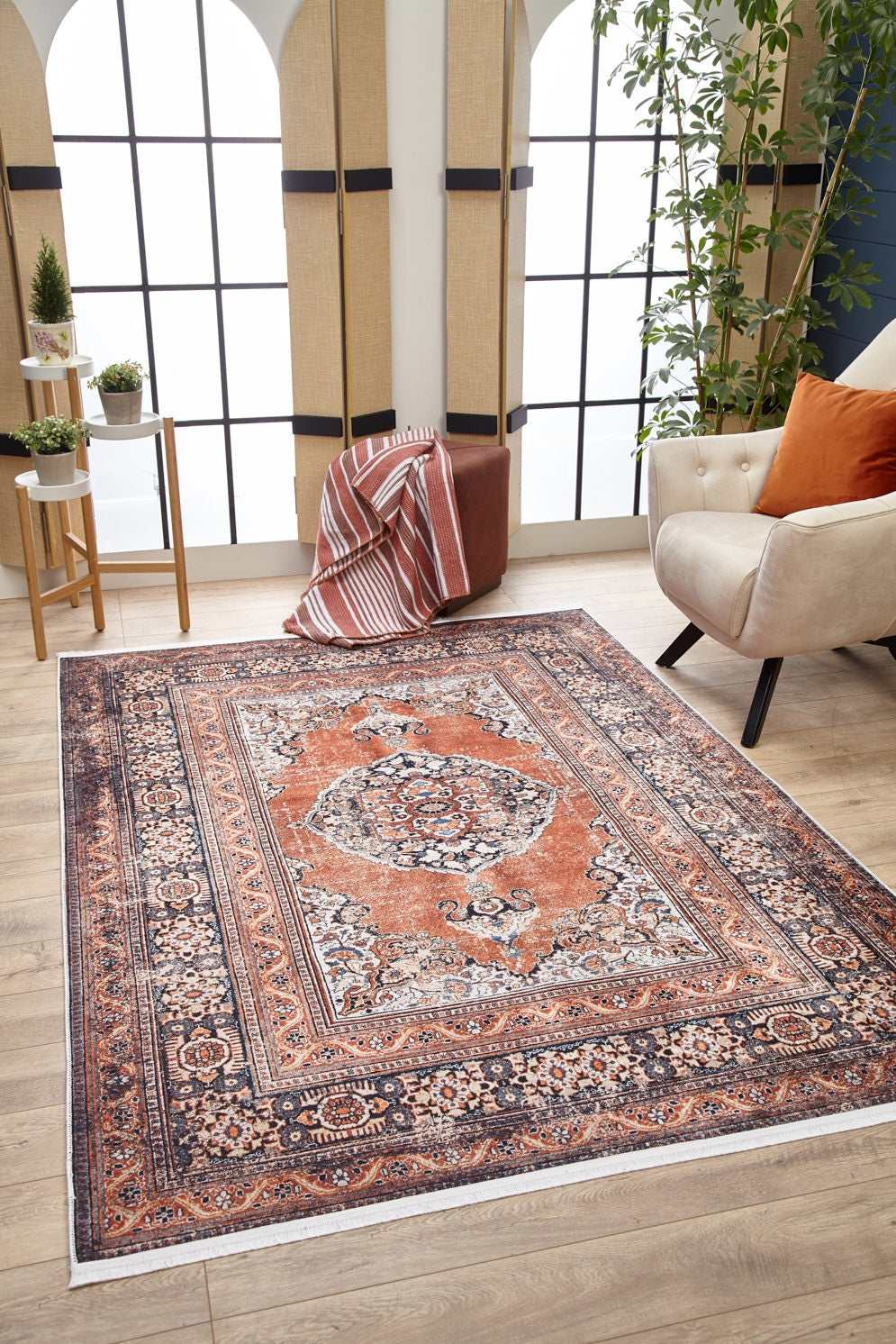 A cozy living room with the Golden Legacy - Washable Rug - JR1931 in orange and navy, ideal for high-traffic areas, a cream armchair with rust cushion, a small round table with plants, a pouf draped with a striped blanket, and large arched windows.