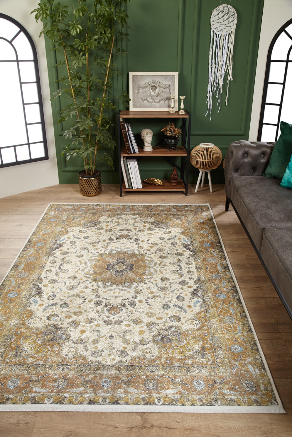 A living room with the Golden Oasis - Washable Rug - JR1930 on wooden floors—perfect for high-traffic areas—features a green wall, gray sofa with green pillows, small decorated bookshelf, potted plant, and a dreamcatcher on the wall.