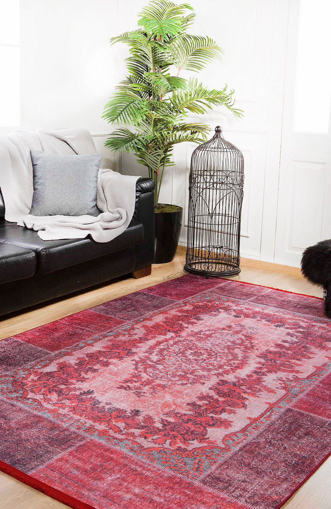 A living room with a black leather sofa, gray blanket and pillow, potted plant, large birdcage, and the Golden Wonder - Washable Rug - JR306 in red—perfect for high-traffic areas—on a light wooden floor.