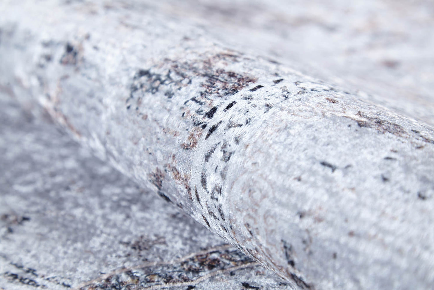A close-up of the Heritage Jewel - Washable Rug - JR1778 showcases its textured, light gray surface with mottled, stone-like patterns in darker shades, reminiscent of a flat weave rug.