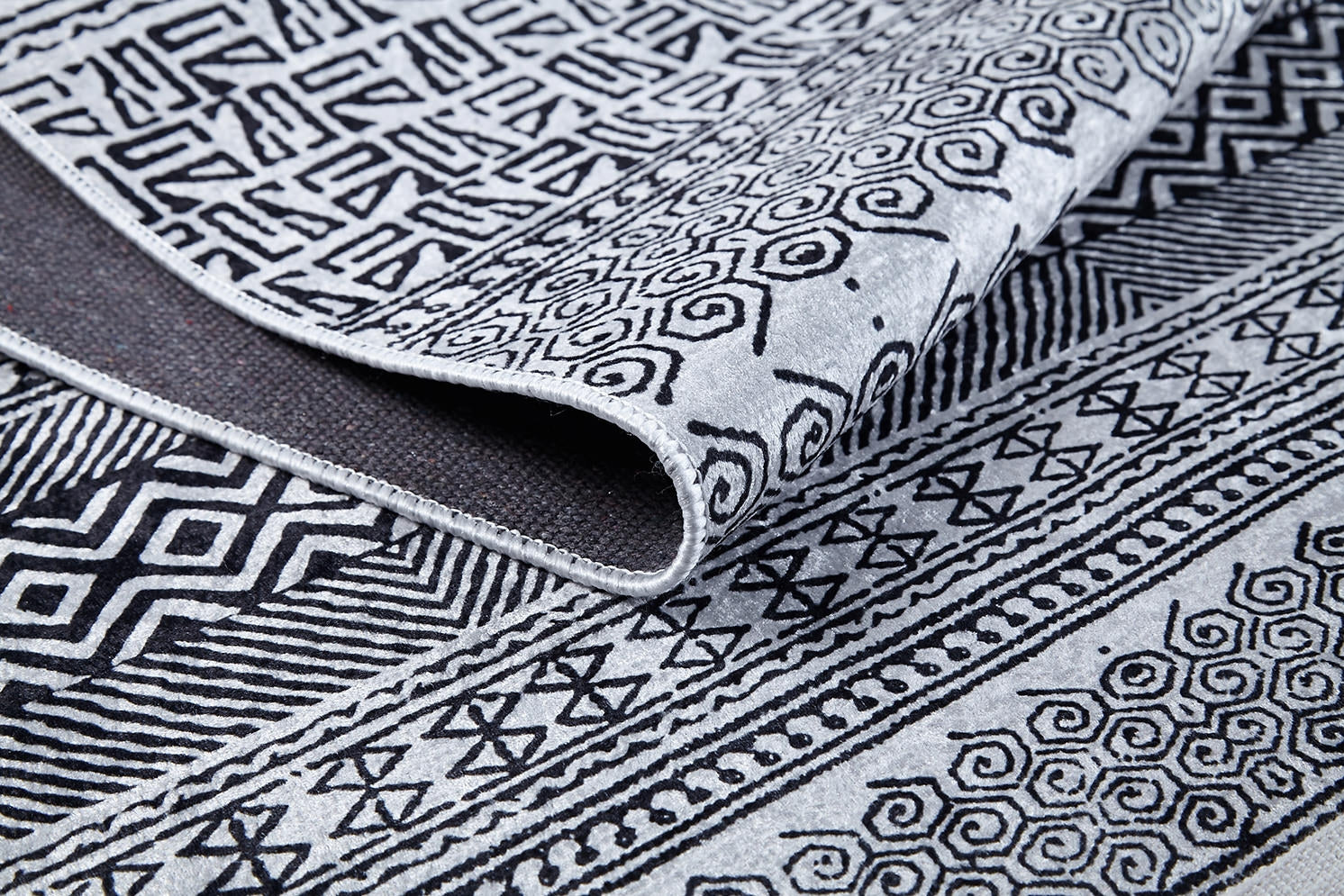 A close-up of the Indie Impressions - Washable Rug - JR1648 showcases its black and white geometric and swirl patterns, with the corner of the monochrome rug slightly folded to display its washable design.