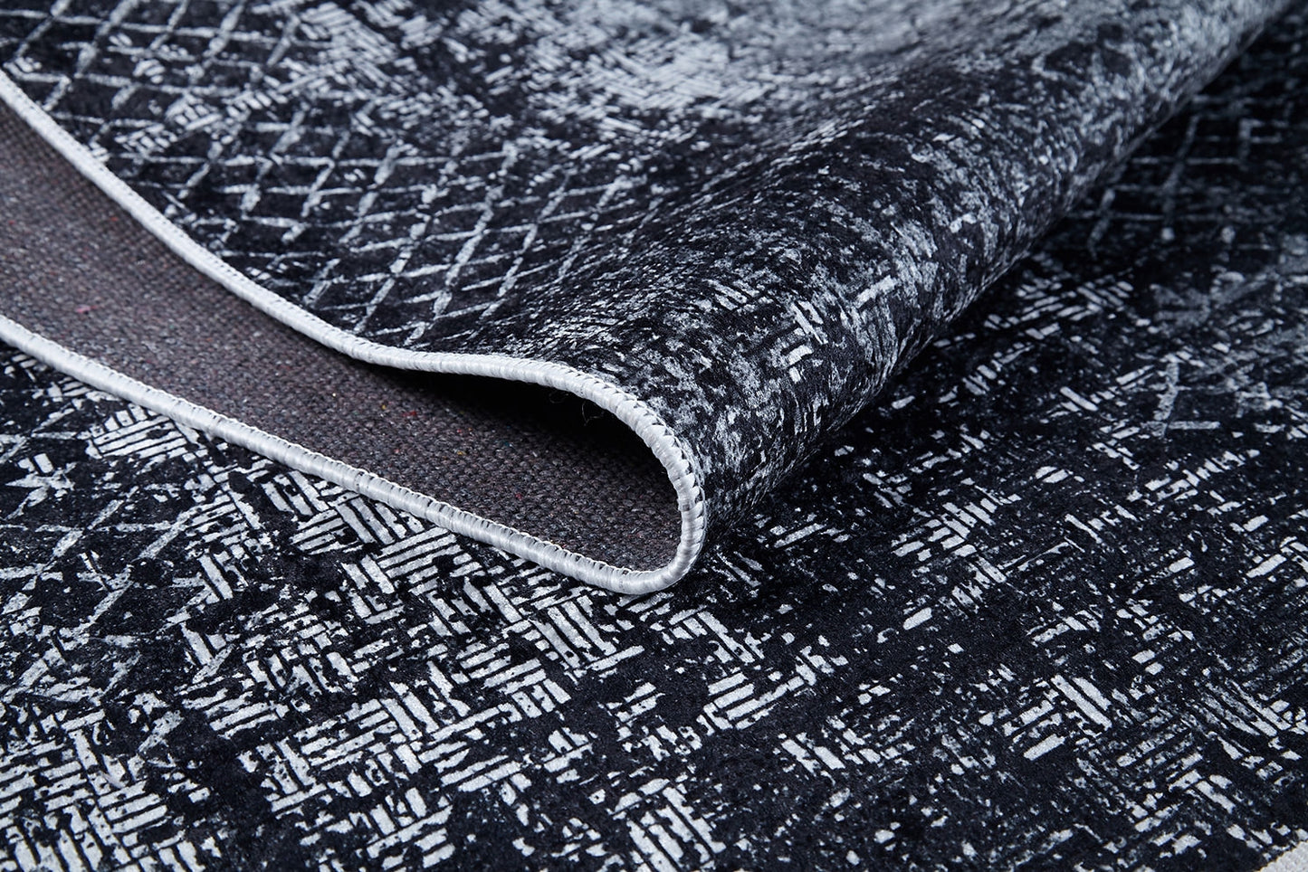 A close-up of the Infinity Garden - Washable Rug - JR1749 shows its black and white abstract geometric pattern, textured surface, lifted edge to reveal the underside and border stitching—durable and ideal for high-traffic areas.