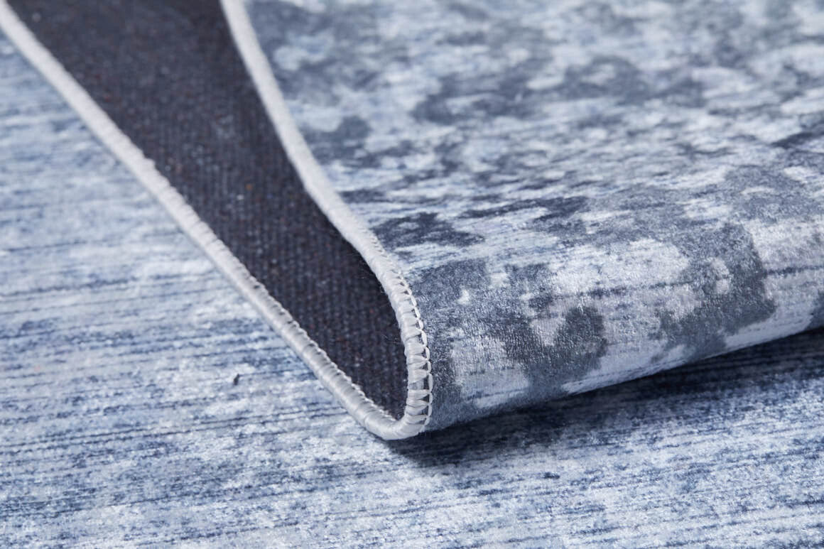 A close-up of the Intricate Scroll - Washable Rug - JR342 shows its stitched edge and textured backing. The mottled blue and gray abstract pattern is perfect for high-traffic areas, bringing modern style to your space.