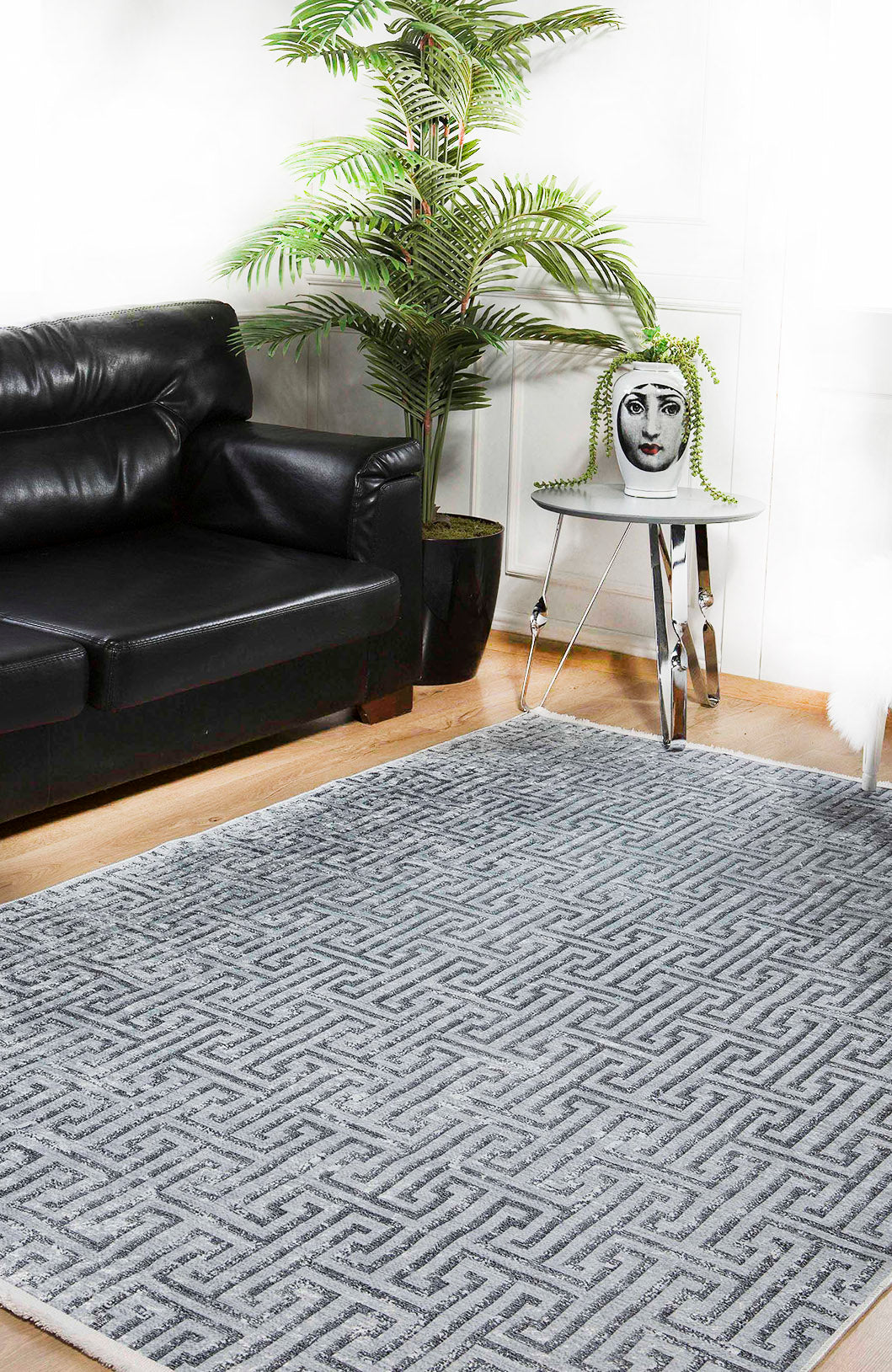 A modern living room with a black leather sofa, large potted plant, metallic side table with a face-printed vase, and the Jazz Age - Washable Rug - JR1225 in gray-anthracite—ideal for high-traffic areas—on a wooden floor.