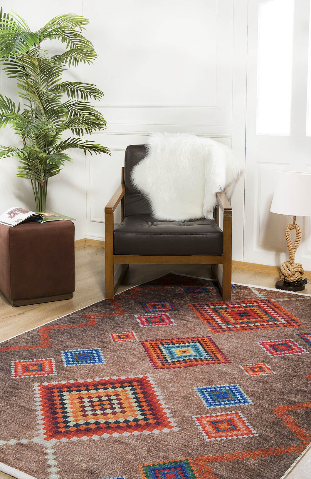 A cozy reading nook with a brown armchair draped in a white faux fur throw, the Kilim Kingdom Washable Rug (JR1536), a small ottoman holding a magazine, a potted plant, and a table lamp on light wooden floors.