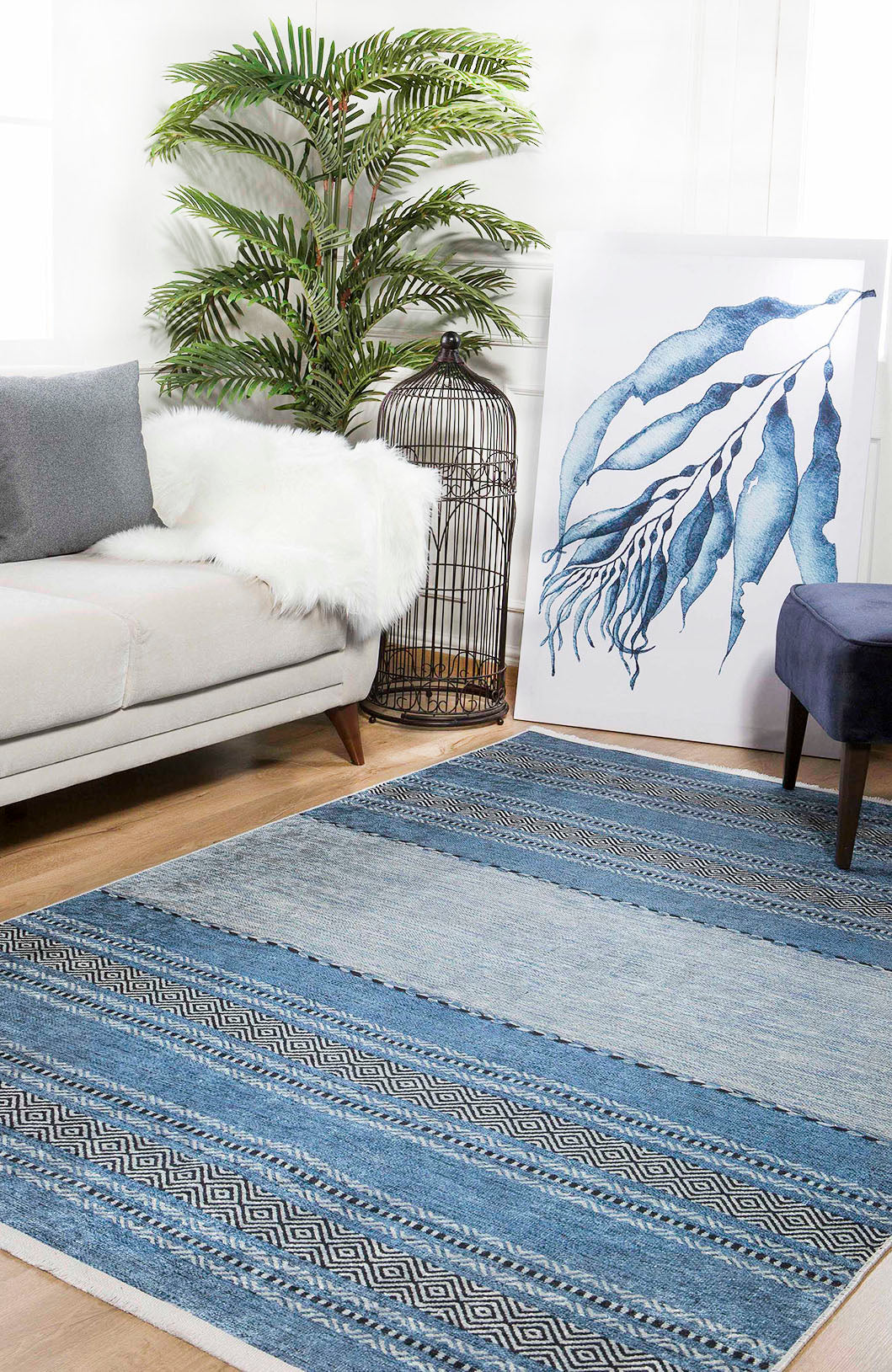 A modern living room featuring the Layered Lines - Washable Rug - JR1545 in blue patterns, ideal for high-traffic areas, paired with a light gray sofa, white faux fur throw, potted plant, birdcage, blue artwork, and dark blue stool on wood flooring.
