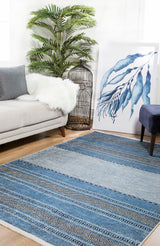 A modern living room featuring the Layered Lines - Washable Rug - JR1545 in blue patterns, ideal for high-traffic areas, paired with a light gray sofa, white faux fur throw, potted plant, birdcage, blue artwork, and dark blue stool on wood flooring.