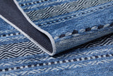 A close-up of the Layered Lines - Washable Rug - JR1545 shows its geometric blue patterns, a slightly folded edge revealing the underside and stitching—ideal for high-traffic areas.