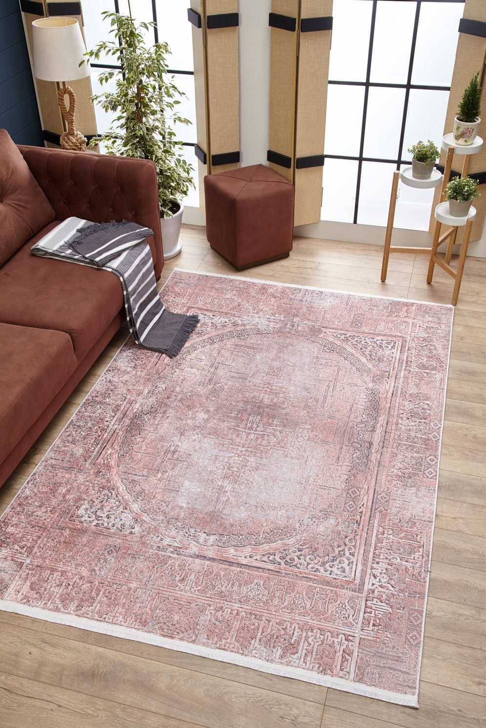 A living room with a brown sofa, striped throw, Luxurious Design - Washable Rug - JR1924 in soft pink—ideal for high-traffic areas—potted plants, a brown ottoman, side table, and large windows letting in natural light.