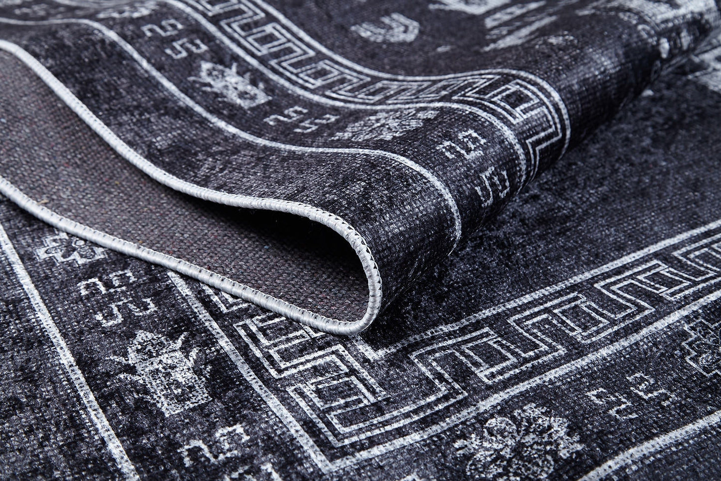 A close-up of the Majestic Symbol - Washable Rug (JR1892) in a dark tone, featuring intricate white geometric and floral designs, highlights its flat weave texture and detailed edge stitching with part of the rug folded over.