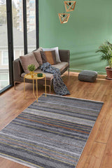 A cozy living room features a small beige sofa, patterned throw blanket, yellow side table, Metro Lines - Washable Rug - JR1548 in gray-anthracite for high-traffic areas, potted plant, floor cushion, geometric hanging lights, green walls, and large windows.