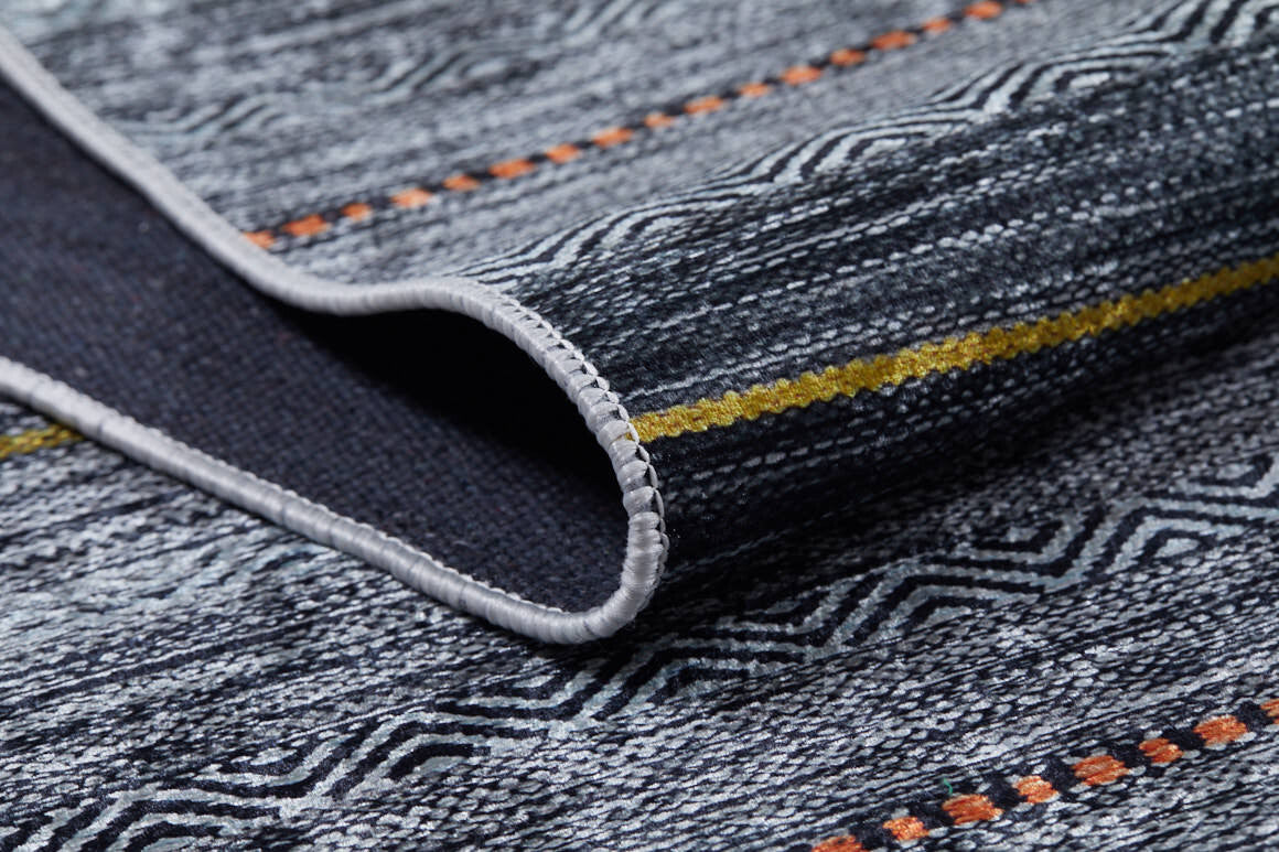 A close-up of the Metro Lines - Washable Rug - JR1548 shows its textured gray-anthracite weave, black geometric patterns, yellow and orange lines, and neatly stitched edges. A folded corner reveals the washable underside—ideal for high-traffic areas.