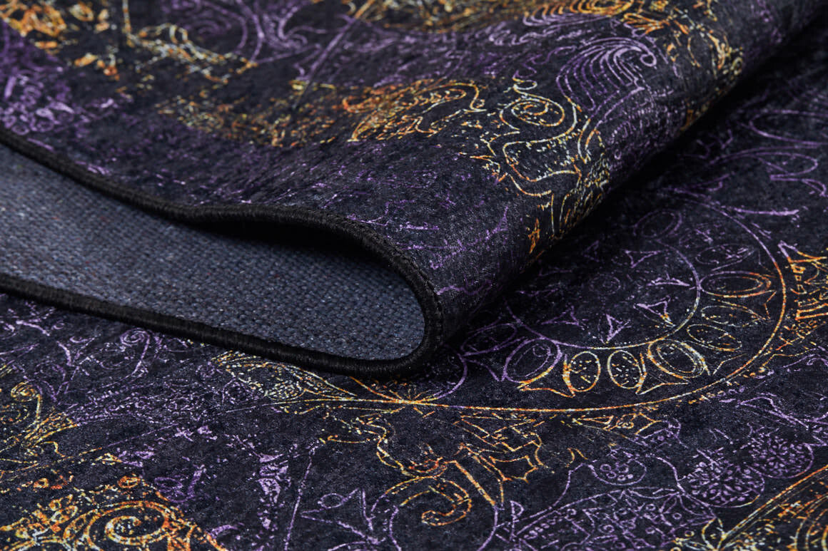 A close-up of the Minimalist and Elegant - Washable Rug - JR1298 reveals dark fabric with gold, purple, and orange patterns. Its swirling designs suit high-traffic areas, and a fold shows the textured underside.