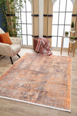 A living room with large grid windows, the Modern Mosaic - Machine-Washable Rug JR1846 in orange and gray for high-traffic areas, a beige armchair with orange pillows, brown ottoman with striped blanket, potted plants, and wood flooring.