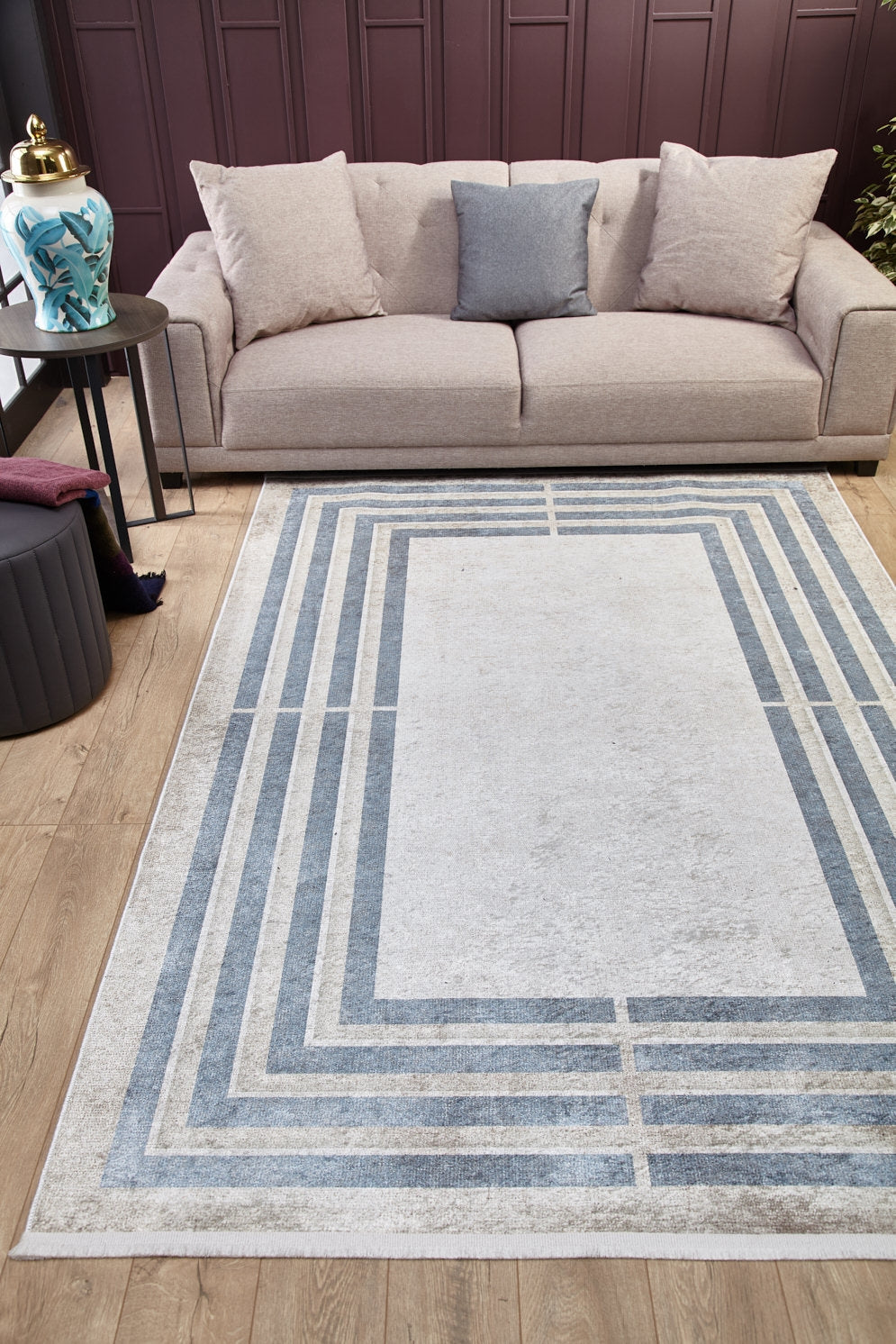 A modern living room with a light beige sofa, three cushions, a decorative vase on a side table, and the Modern and Regal - Washable Rug - JR1616 showcasing blue and gray rectangles on the wooden floor.