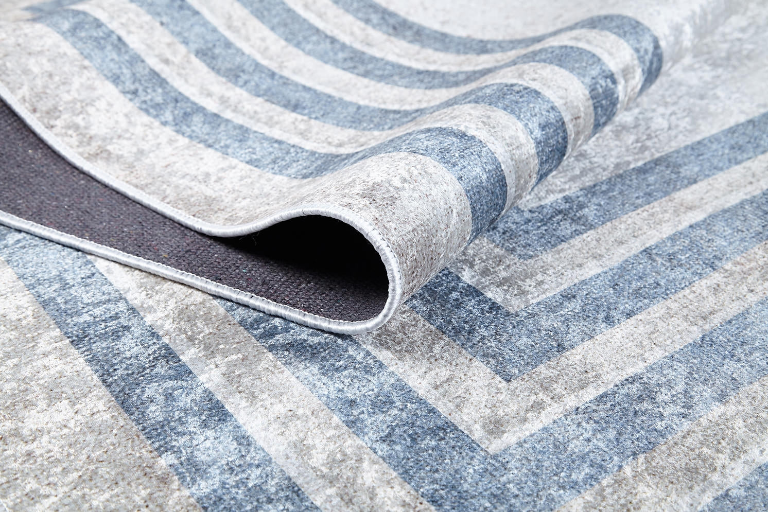 A close-up of the Modern and Regal - Washable Rug - JR1616 shows its blue and gray pattern, with one corner folded to reveal the dark underside and textured fabric.
