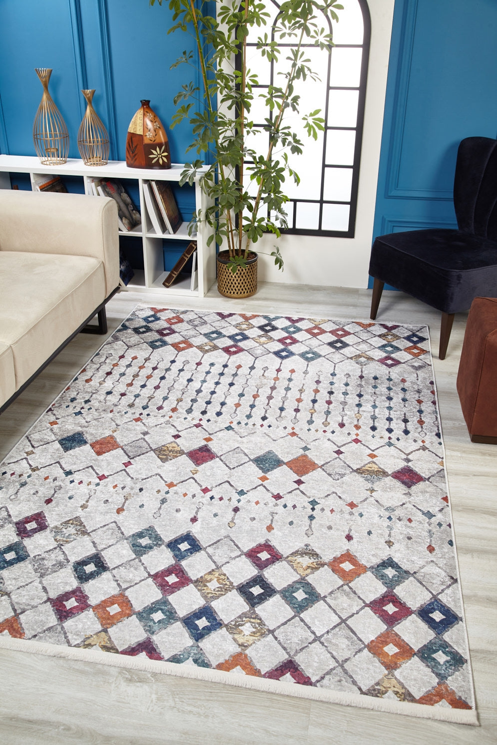 A modern living room features the Moroccan Mosaic - Washable Rug - JR1655 with a geometric pattern—perfect for high-traffic areas—a beige sofa, dark chair, brown ottoman, tall plant, blue wall paneling, window, and decor on a white shelf.