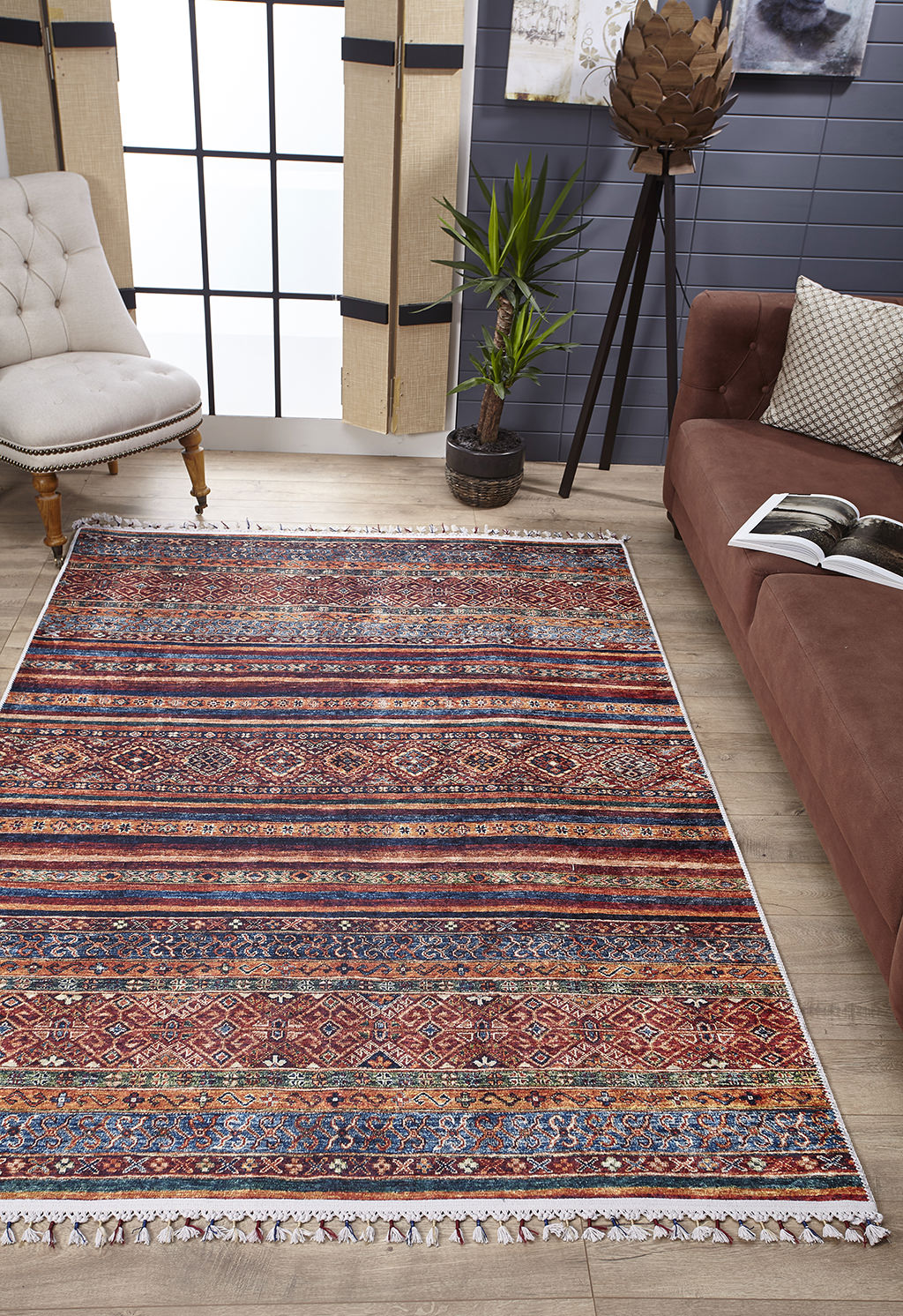 A living room featuring the Native Nightscape - Washable Rug - JR5093, ideal for high-traffic areas, a brown sofa with an open book, a potted plant, a tufted beige chair, large windows with natural light, and light wood floors.