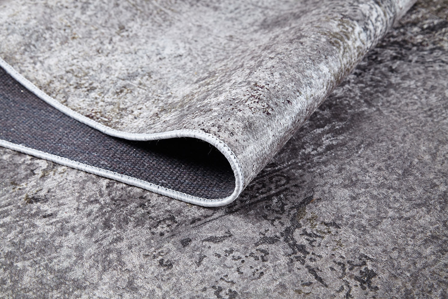 A close-up of the Noble Insignia - Washable Rug - JR1830 shows its gray textured surface with one corner lifted to reveal the black underside, highlighting durable material and stitching—perfect for high-traffic spaces.