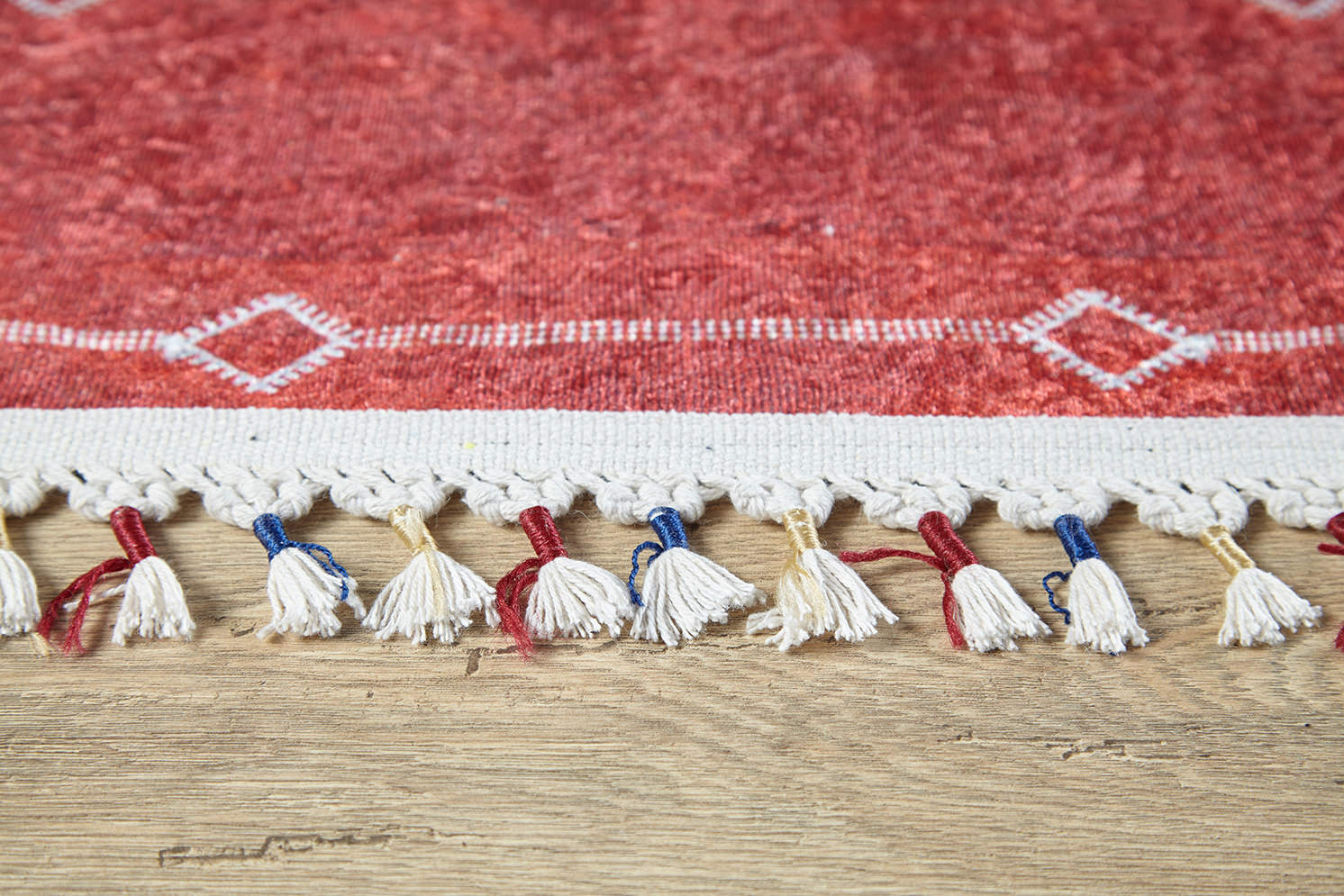 Close-up of the Nomadic Journey - Washable Rug - JR5080: a red rug with white geometric edge designs, white fringe with red and blue threads. Washable and perfect for high-traffic areas, it complements wooden floors beautifully.