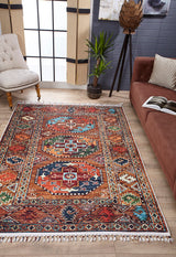 The Nomadic Oasis - Washable Rug - JR5086, featuring colorful geometric patterns, is displayed on a wooden floor in a modern living room with a brown sofa, cream armchair, potted plants, and large windows with sheer beige curtains.