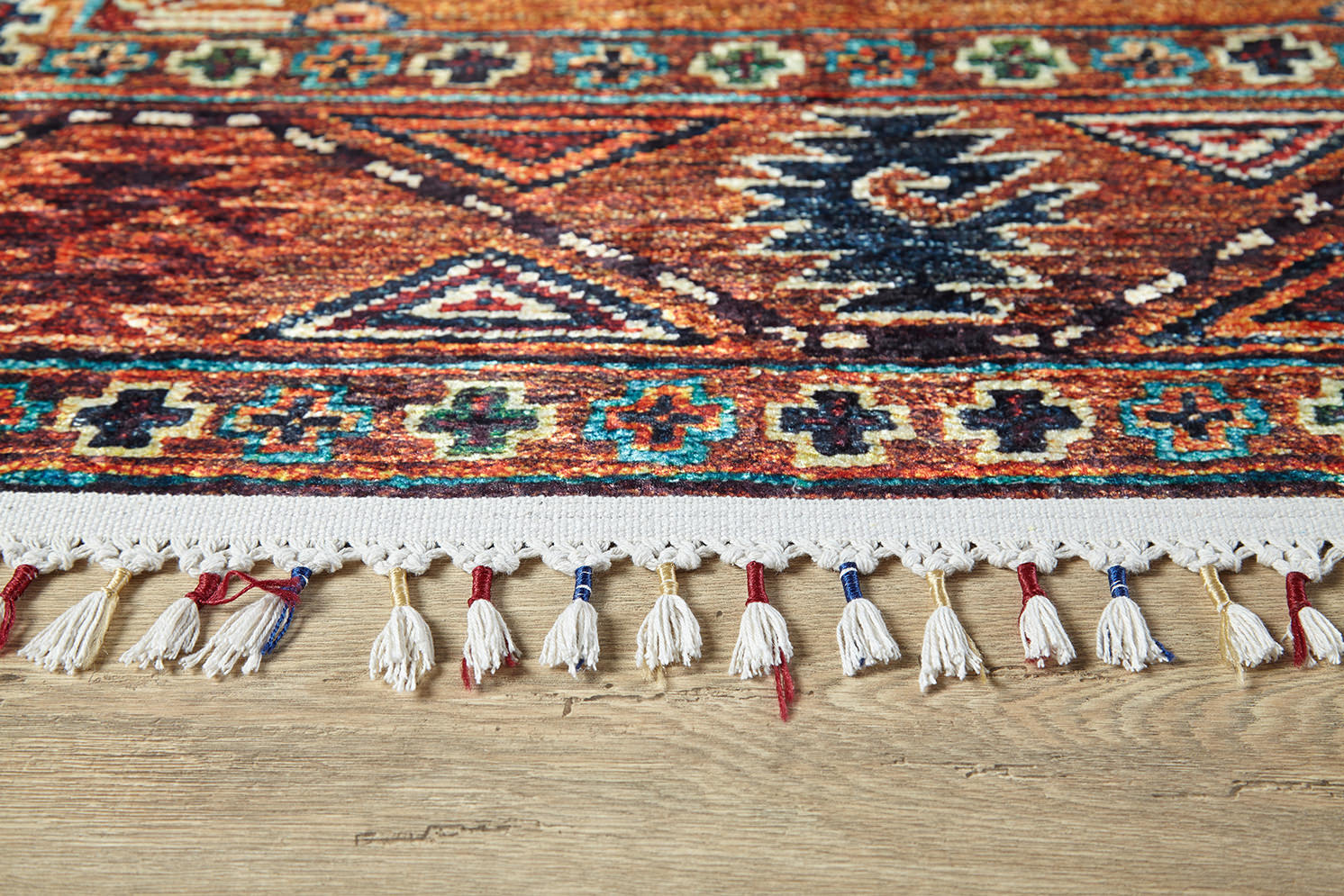 Close-up of the Nomadic Oasis - Washable Rug - JR5086, a colorful flat weave with fringed edges, blue, red, and beige geometric patterns, tassels, and placed on a wooden floor.
