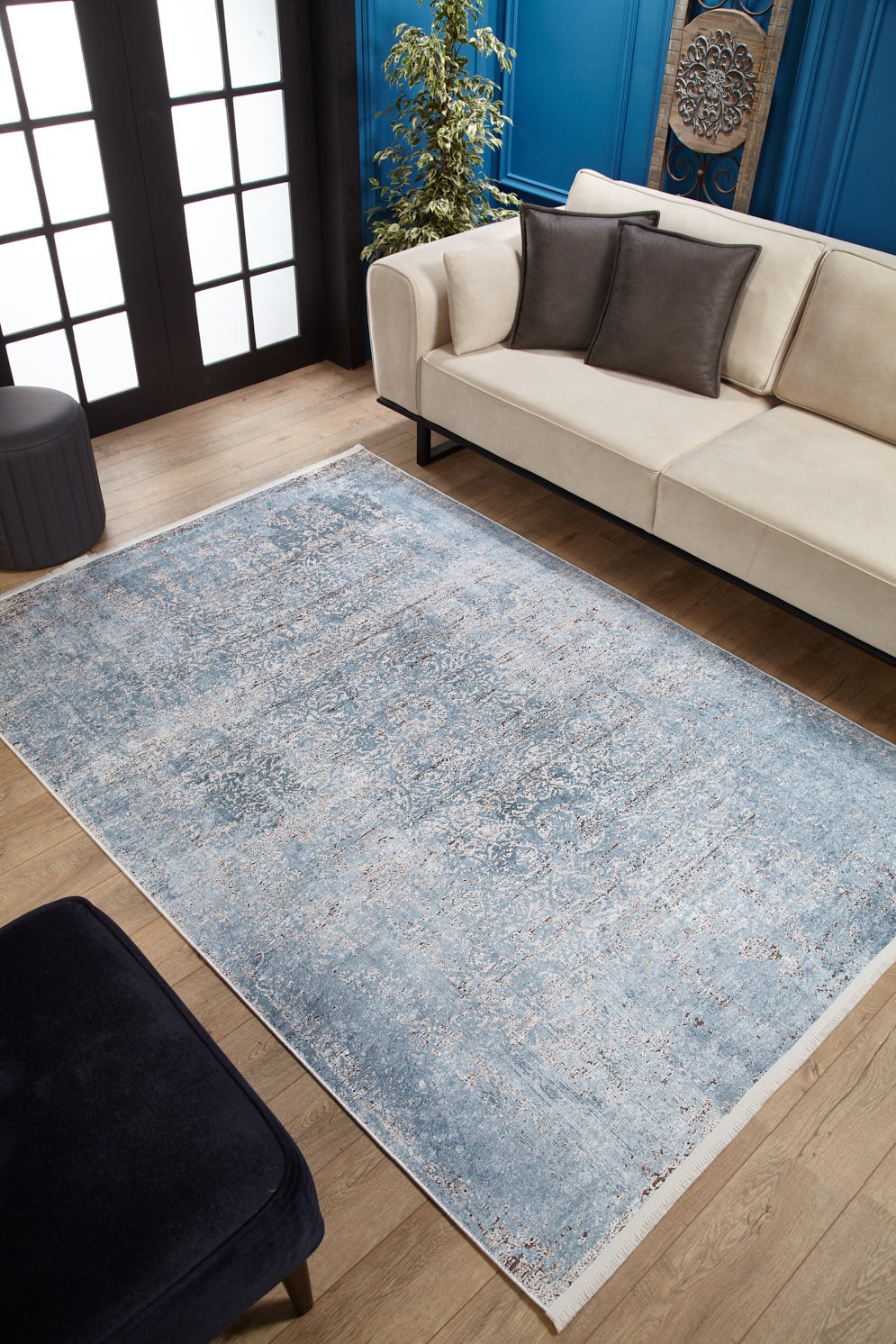 A modern living room features the Old World Charm - Washable Rug - JR1606 in blue on wooden floors, paired with a beige sofa, dark pillows and ottoman, green plant, and accented blue panel walls—perfect for high-traffic spaces.