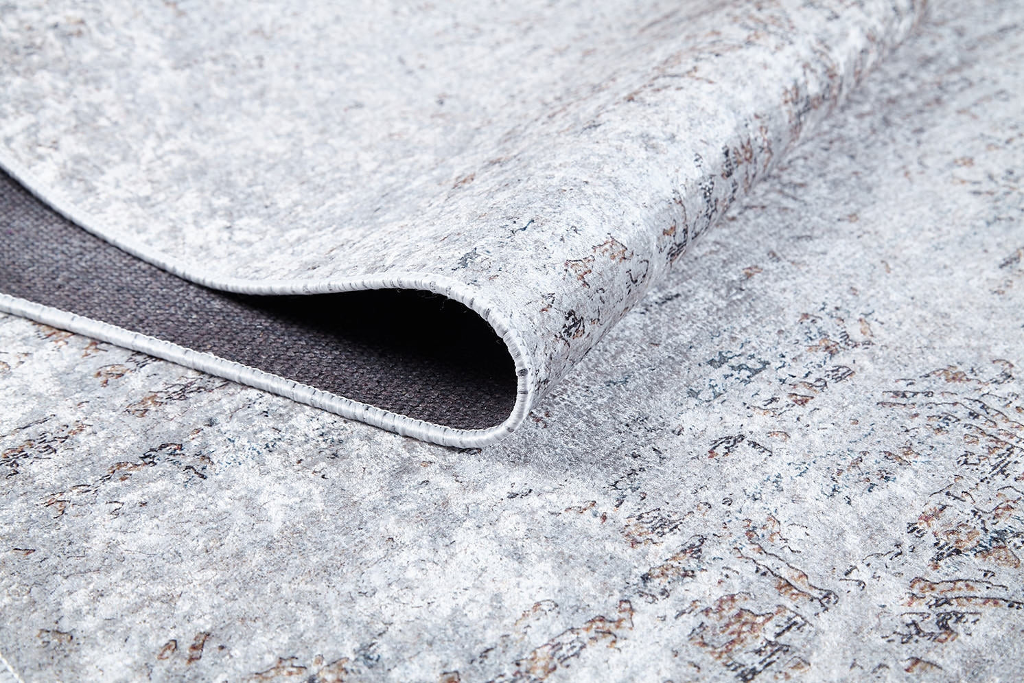 A close-up of the Opulent Tapestry - Washable Rug - JR1775 shows its textured, light gray and brown weathered pattern with a folded corner displaying the dark underside—perfect for high-traffic spaces.