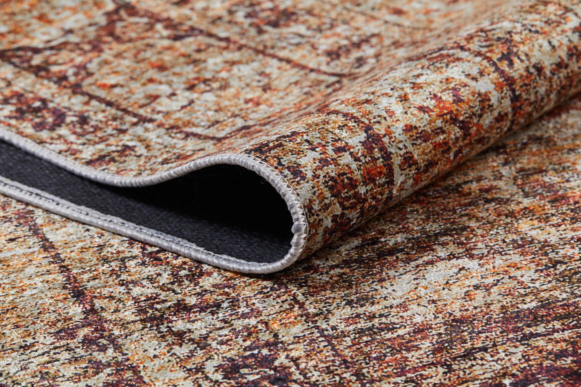 Close-up of a corner of the Ornate Grace - Washable Rug - JR1009, featuring a textured, multicolored surface in brown, orange, and beige. The slightly lifted edge reveals a black underside and stitched border—perfect for high-traffic areas.