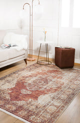 A cozy living room features a beige sofa, brown ottoman, side table with a vase, two tall floor lamps, and the Ornate Grace - Washable Rug - JR1009 in red and beige—perfect for high-traffic areas—on a wooden floor.