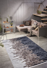 A modern living room features a gray sectional sofa, assorted pillows, light wood floors, and a metal shelf with plants. The space is anchored by the Pixelated Paradise - Machine-Washable Rug JR902 in bronze and brown tones, perfect for allergy-sensitive homes.