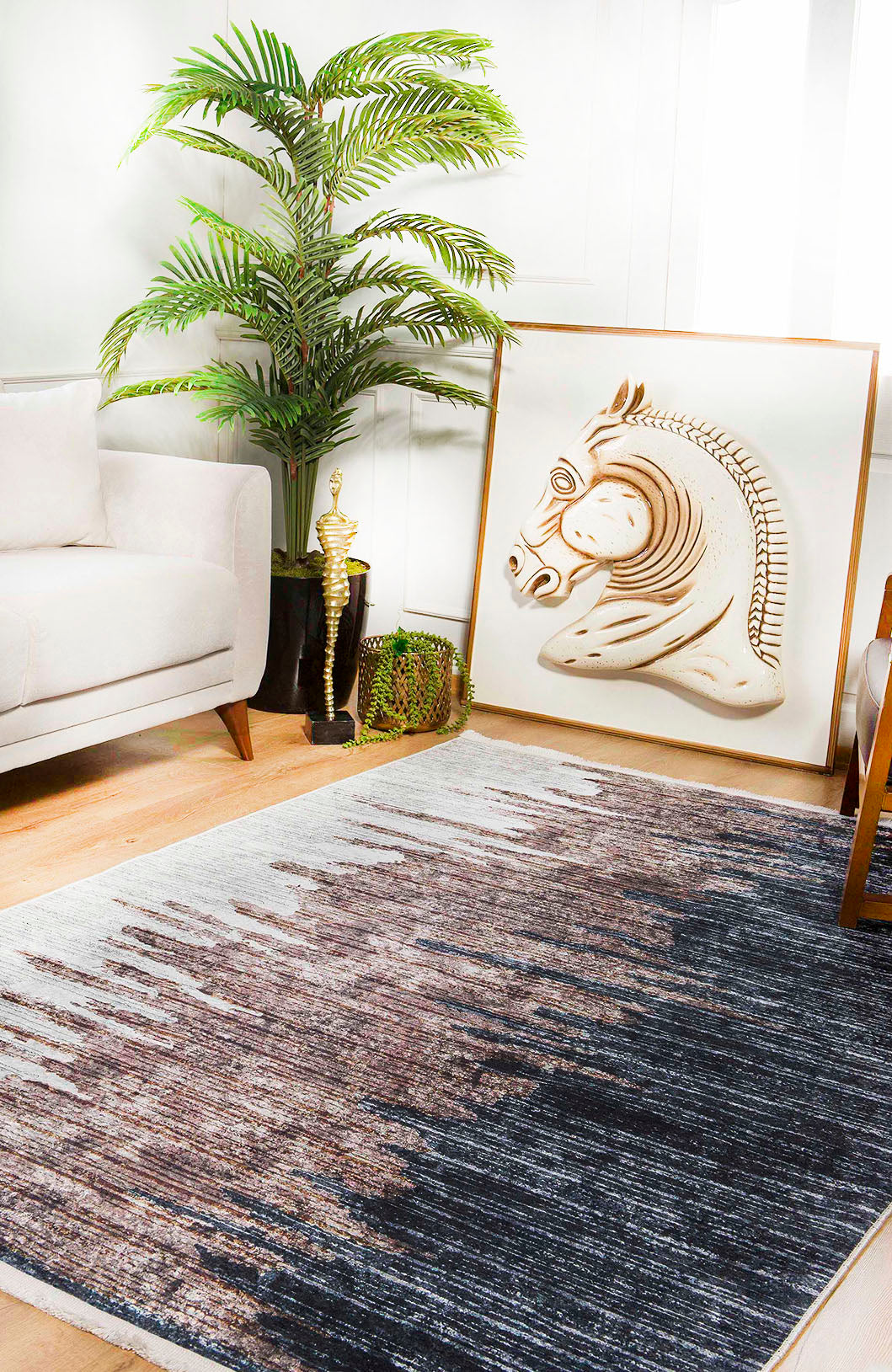 A modern living room features a beige sofa, leafy potted plant, decorative trophy, and large framed horse head artwork on the floor. The wooden floor is covered by the Pixelated Paradise - Machine-Washable Rug JR902 in bronze and brown.