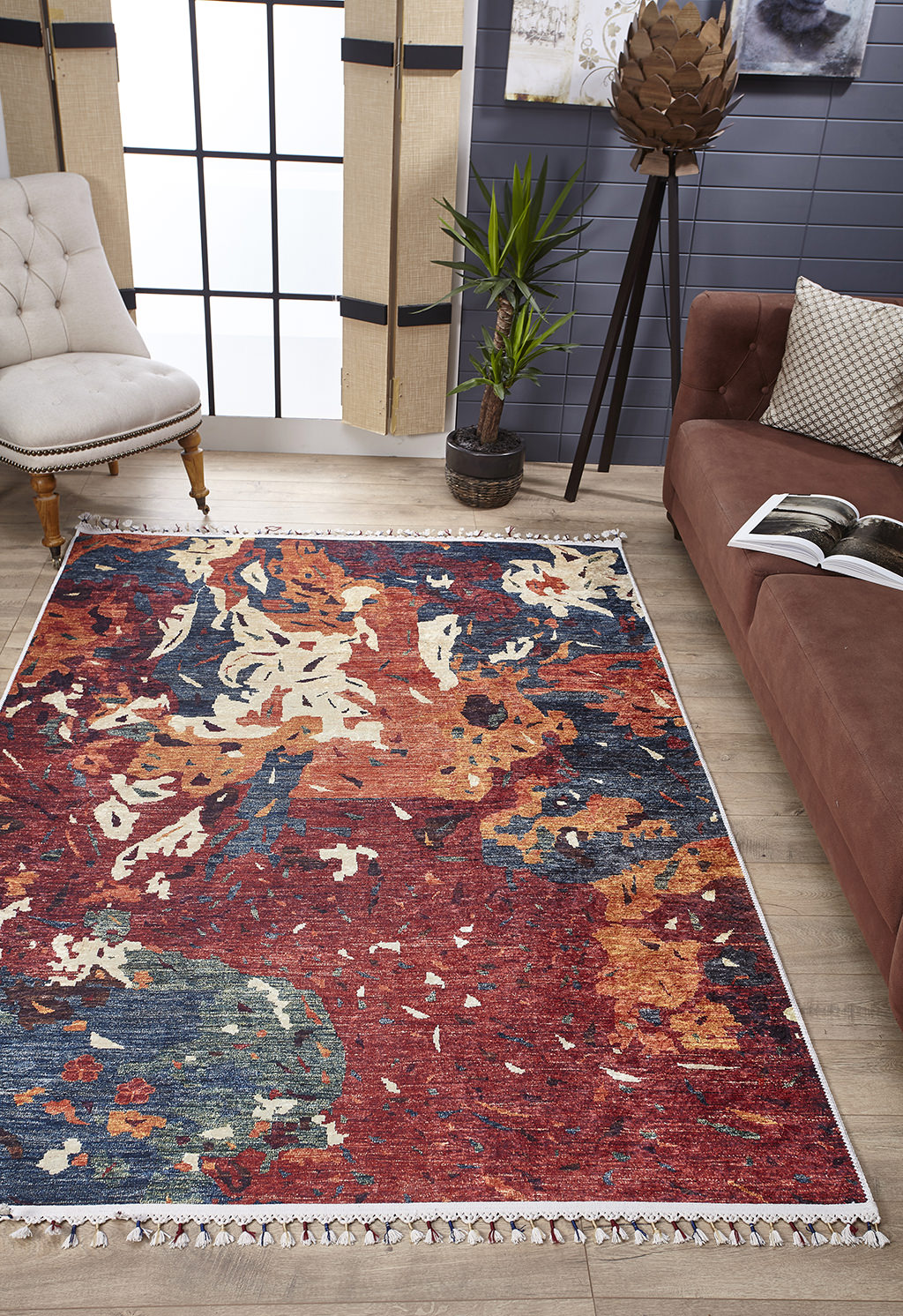 A cozy living room featuring the Psychedelic Splash - Machine-Washable Rug - JR5096, perfect for high-traffic areas, a brown sofa with a patterned cushion, beige tufted chair, potted plant, decorative floor lamp, gray-paneled walls, and large windows.