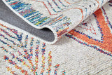 A close-up of the Rainbow Rhapsody - Washable Rug - JR5060 shows a folded corner, highlighting its textured flat weave and vibrant orange, blue, and red geometric designs on a beige background.