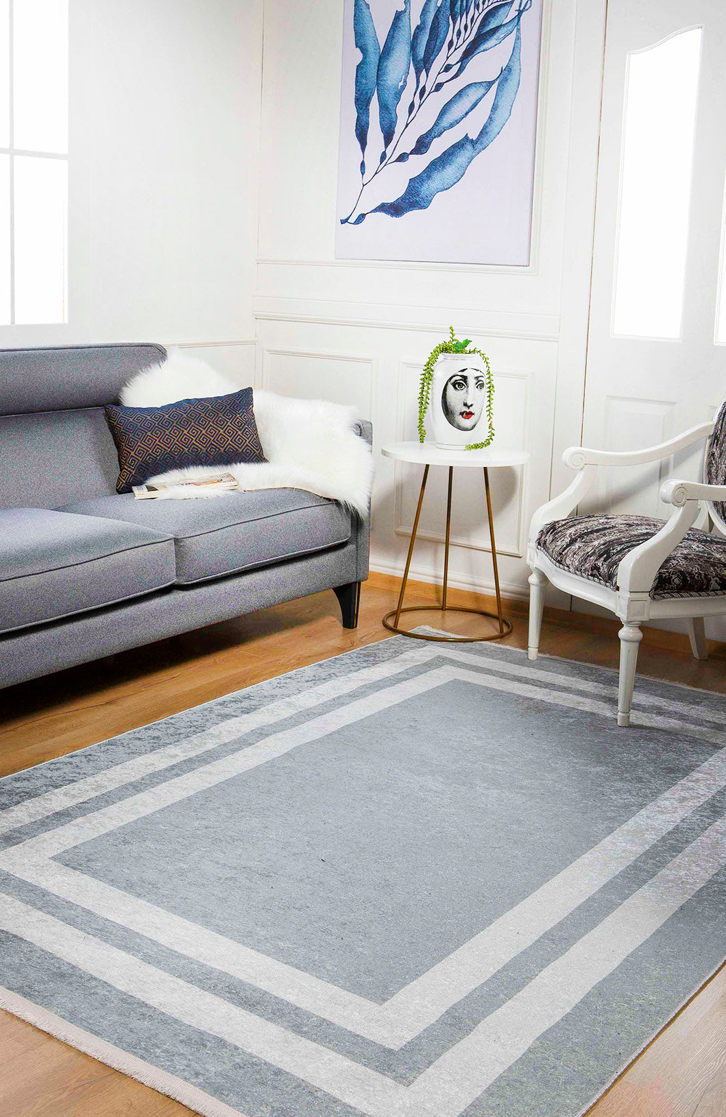 A modern living room with a gray sofa, white armchair, and the Refined Edge - Washable Rug - JR573—ideal for high-traffic spaces. A round side table with a vase of greenery and framed blue leaf artwork complete the look.