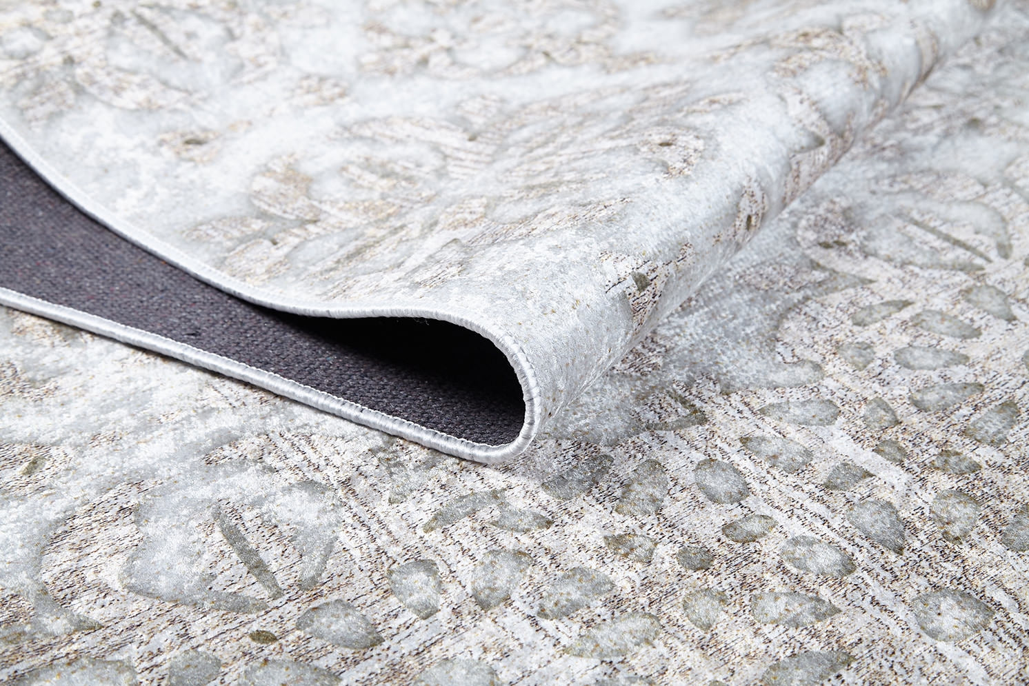 A close-up of the Regal Embellishment - Washable Rug - JR1590 shows a folded corner revealing its dark, textured underside. This washable rug features a subtle beige and gray floral design, ideal for modern interiors.