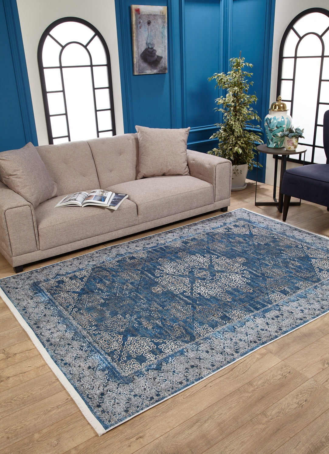 A beige sofa with cushions sits behind an open book, next to the Regal Insignia - Washable Rug - JR1786 on wooden flooring. Blue paneled walls, arched windows, a potted plant, and side tables complete this modern living room for high-traffic areas.