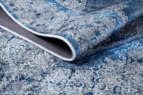 Load image 1 in gallery view. A close-up of the Regal Insignia - Washable Rug - JR1786 in blue, featuring intricate swirling designs and a folded edge revealing the underside—ideal for high-traffic areas.