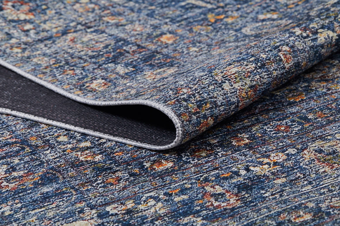 A close-up of the Regal Splendor - Washable Rug - JR1017 shows its blue, multicolored abstract pattern with yellow, orange, and white accents. A folded corner reveals the dark non-slip backing on this textured rug, ideal for high-traffic areas.